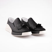 A detailed USA studio shot of the MrCalvano Newport Beach black leather comfort slip-on shoes, highlighting the textured perforated leather, the distinctive tassel and fringe detail on the vamp, and the ergonomic white sole with its contrast stitching. The image emphasizes the clean lines and meticulous construction of the footwear. 