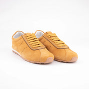 A close-up view of the MrCalvano Pismo Tan Leather Women's Sneaker, showcasing its fine suede texture, flat mustard yellow laces, and subtle serrated trim details. The sole reveals a thin white midsole and a terracotta rubber outsole. Photographed on a clean white background, highlighting the shoe's meticulous construction and warm, inviting hue, perfect for California fashion.